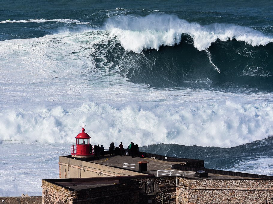 A group of people gathered next to a red lighthouse, watching the giant waves of Nazaré.