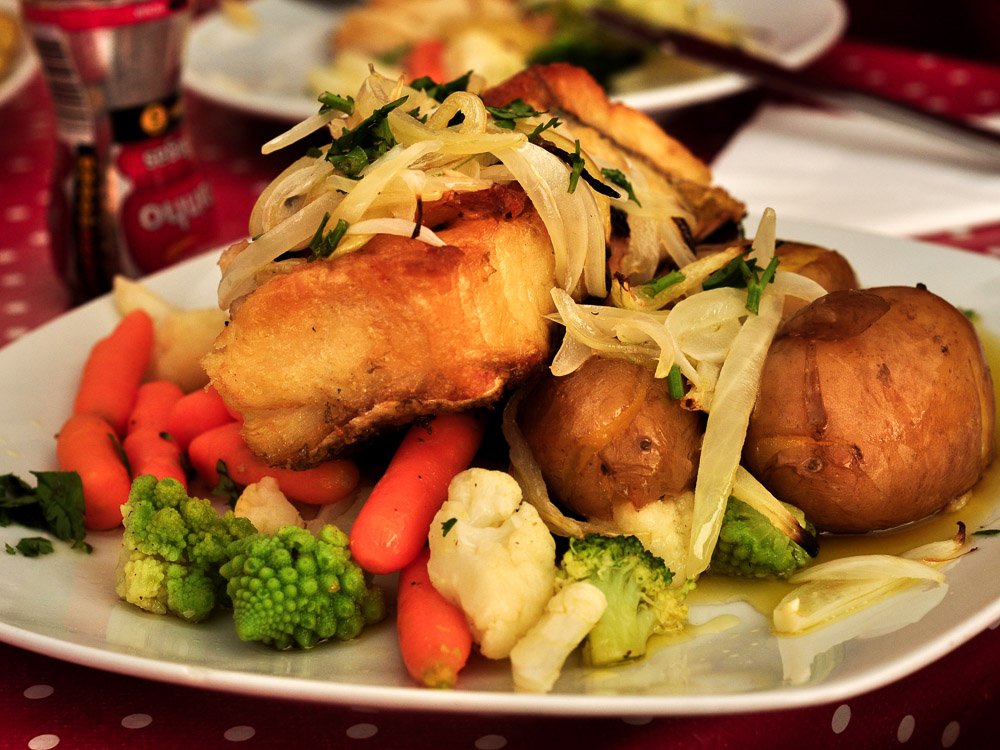 A white plate on top of a red polka-dot tablecloth. A typical Portuguese dish is being served, with codfish, potatoes, carrots, broccoli, and onion.