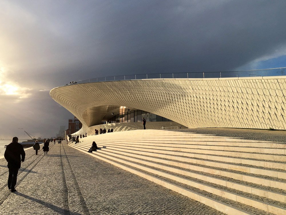 Facade of the MAAT in Lisbon in December. The sky is gray, and a piece of the sun is visible. There is a staircase where people are sitting. There are people walking on the cobblestone sidewalk.