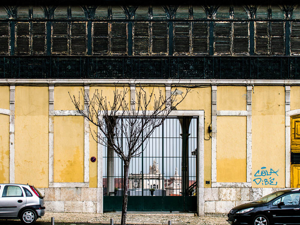 The facade of a building, with a black gate in the middle and a leafless tree in front. The bottom part of the building is yellow, and the top part is black, with old windows. There is a car parked on each side of the gate.