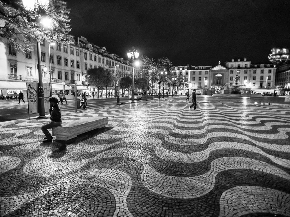 Black-and-white image of a square in Lisbon in December. The ground has black and white wave patterns. There is a person sitting on a bench on the left, and some people walking in the center. There are historic buildings around the square and a fountain in the background.