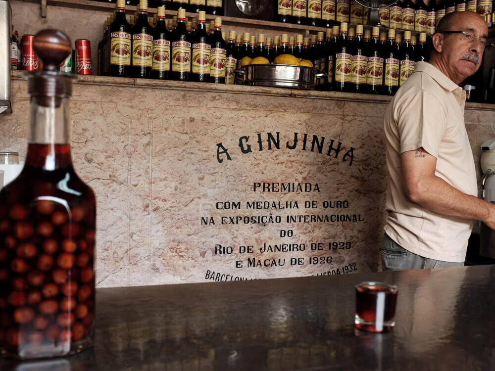 On the right side, a man with glasses and a mustache wearing a beige shirt. Behind him, it says "A Ginjinha" in black letters on a beige wall. Above, there are two shelves full of drinks. In front, a bottle of ginja and a shot glass.