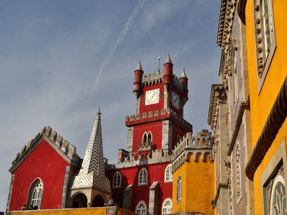 Part of Pena Palace with a blue sky with some clouds in the background. On the left side, a clock tower with red walls and a white tower with black patterns below. On the right side, the construction continues, but with yellow walls.