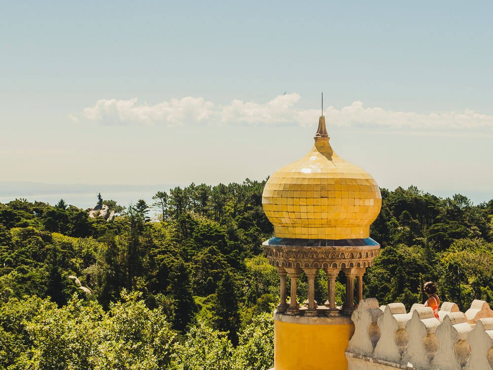 A yellow tower, with columns in the middle, against a backdrop of vegetation and a clear sky.