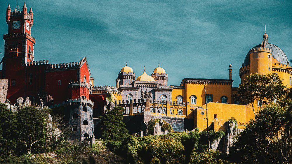 View of Pena Palace. On the left, the part painted red, in the center the part with tiles, and on the right the yellow part. At the bottom, we see vegetation surrounding the castle.