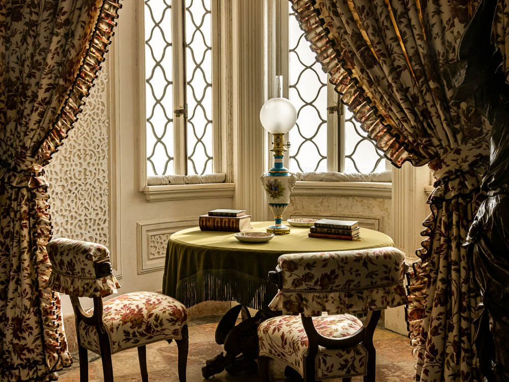 Room in Pena Palace, with ornate windows and flowery curtains. In the center, a table with two chairs upholstered in the same pattern as the curtains. There are books and a lamp on the table.