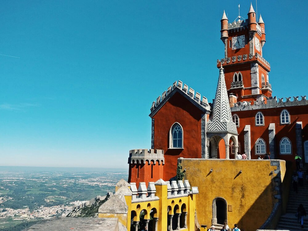 Pena Palace, with its red clock tower and part of the yellow construction. In the background, the clear blue sky, overlooking the city below.