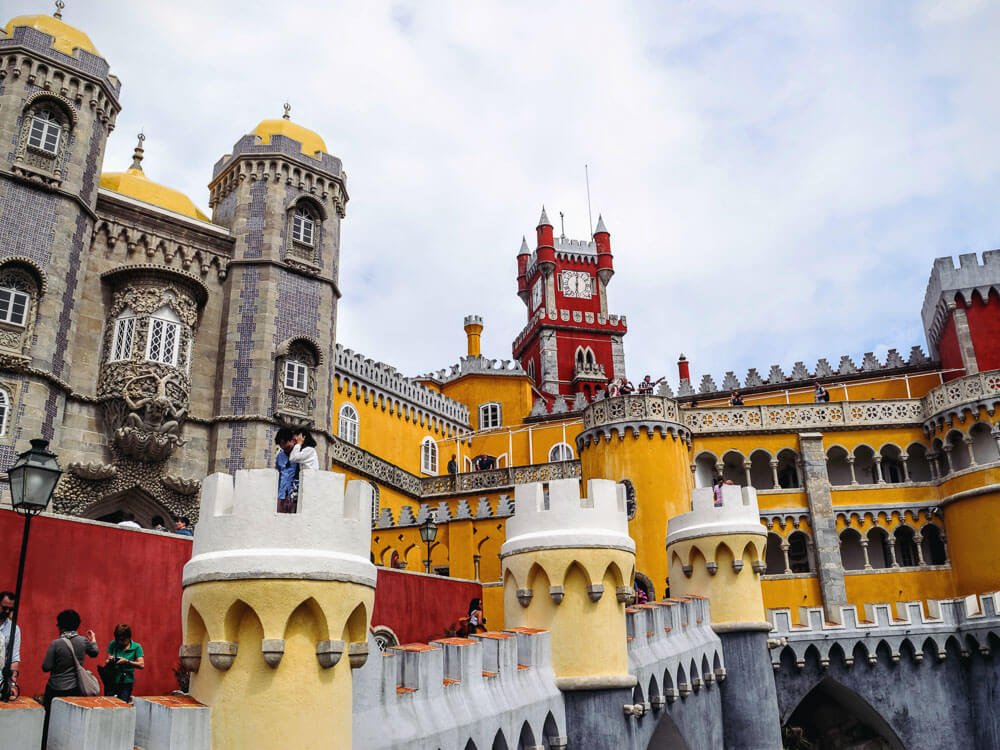 Pena Palace, with colorful towers in red and yellow. On the left side, a building with tiles. There are people strolling around.