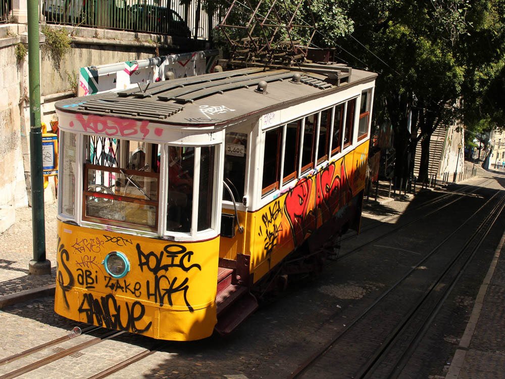 Elevador da Bica ascending the street. There are graffiti on the yellow-painted walls, and in the background, a tree.