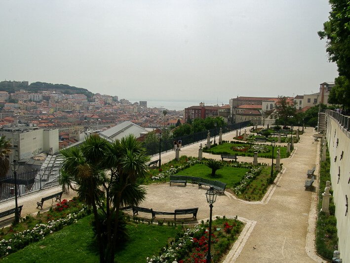 Miradouro de São Pedro de Alcântara, with five flower beds with grass and flowers, and a concrete path passing between them. On the left side, a view of the city of Lisbon.