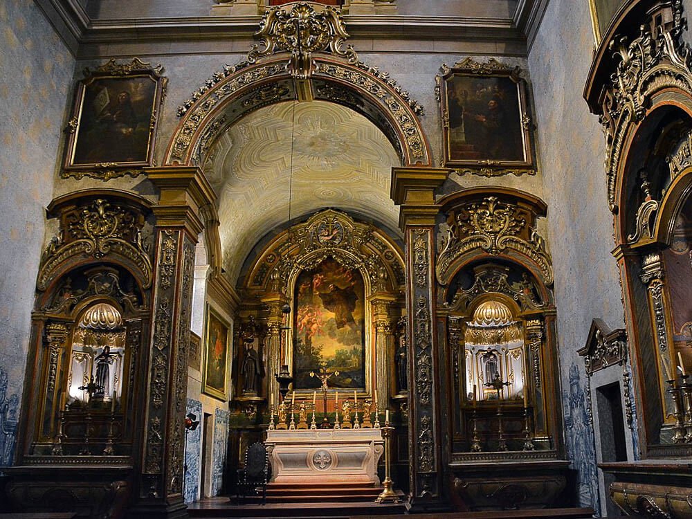 Interior of the Convent of São Pedro de Alcântara. Portuguese tiles adorn the walls, as well as paintings and golden arches. The altar is adorned with candles, and there is a large painting behind it framed with a golden arch.