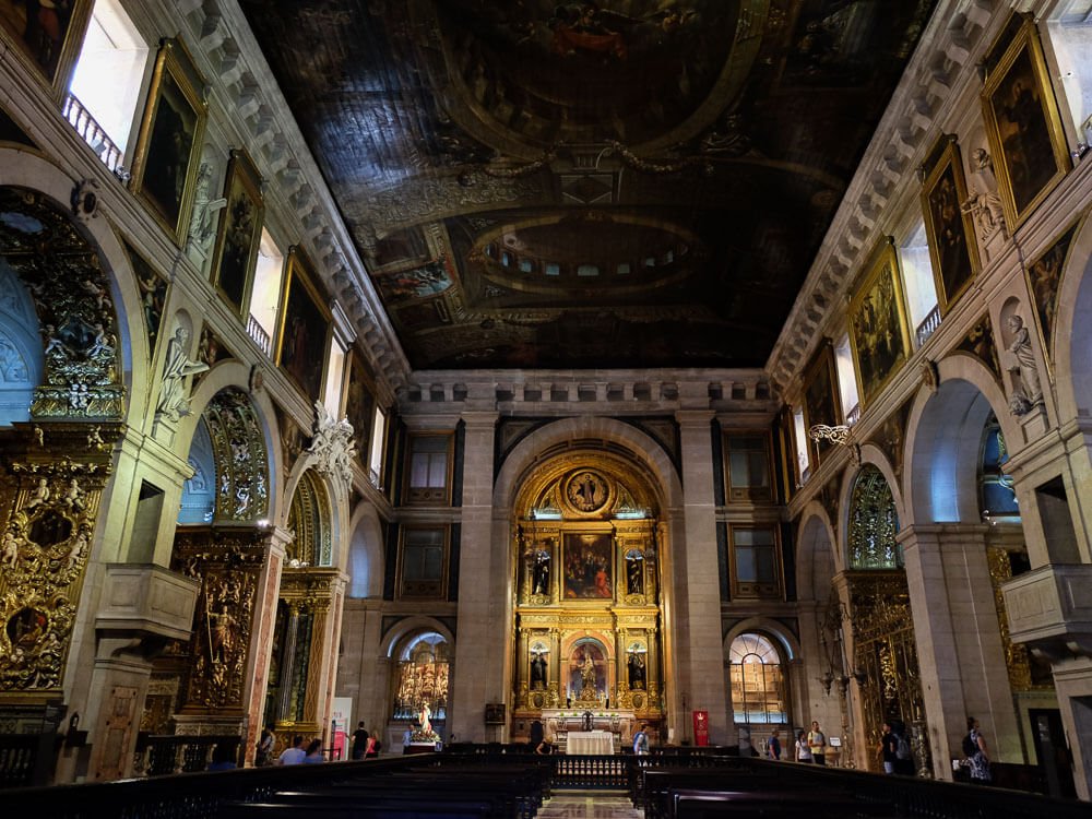 Interior of the Church of São Roque. The ceiling is painted, there are arches with golden details, and a golden altar with images of saints. There are people visiting the church.