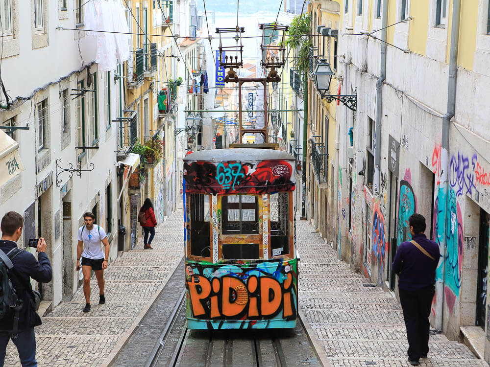 Elevador da Bica ascending the street. There are four people walking up or down the street. The walls and the tram have colorful graffiti.