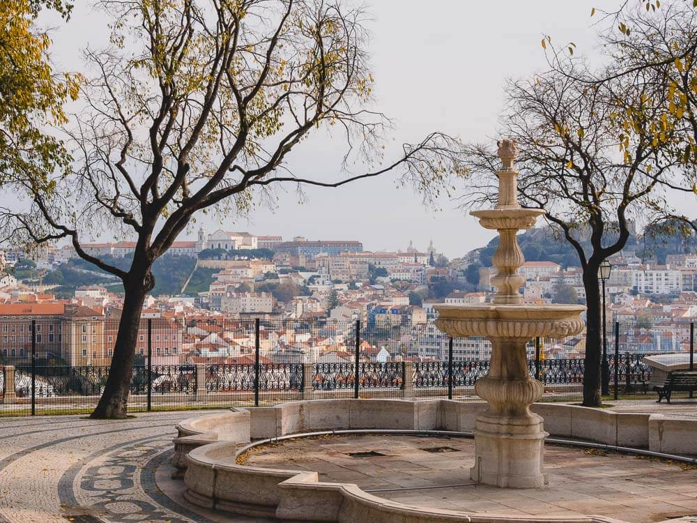 In the foreground, a dry fountain with two sparsely leaved trees. Behind it, a black fence, and in the background, a view of Lisbon.