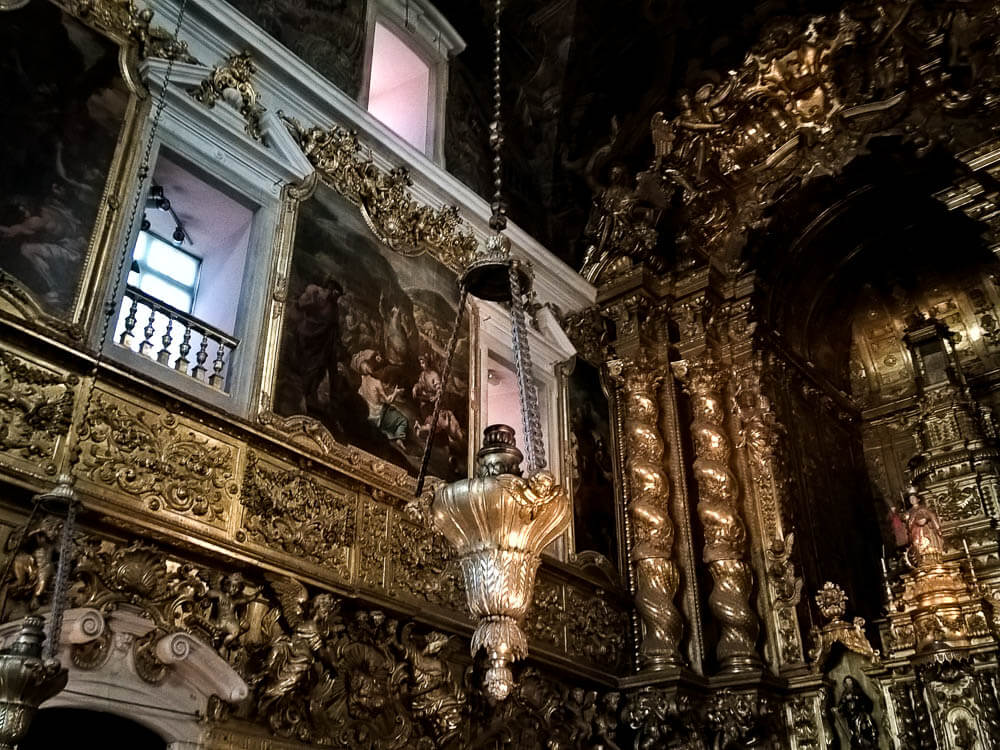 Interior of the Church of Santa Catarina. On the walls, there are paintings with golden frames and other decorations in gold. In the center, a chandelier. On the right side, an all-gold altar with rococo decorations.