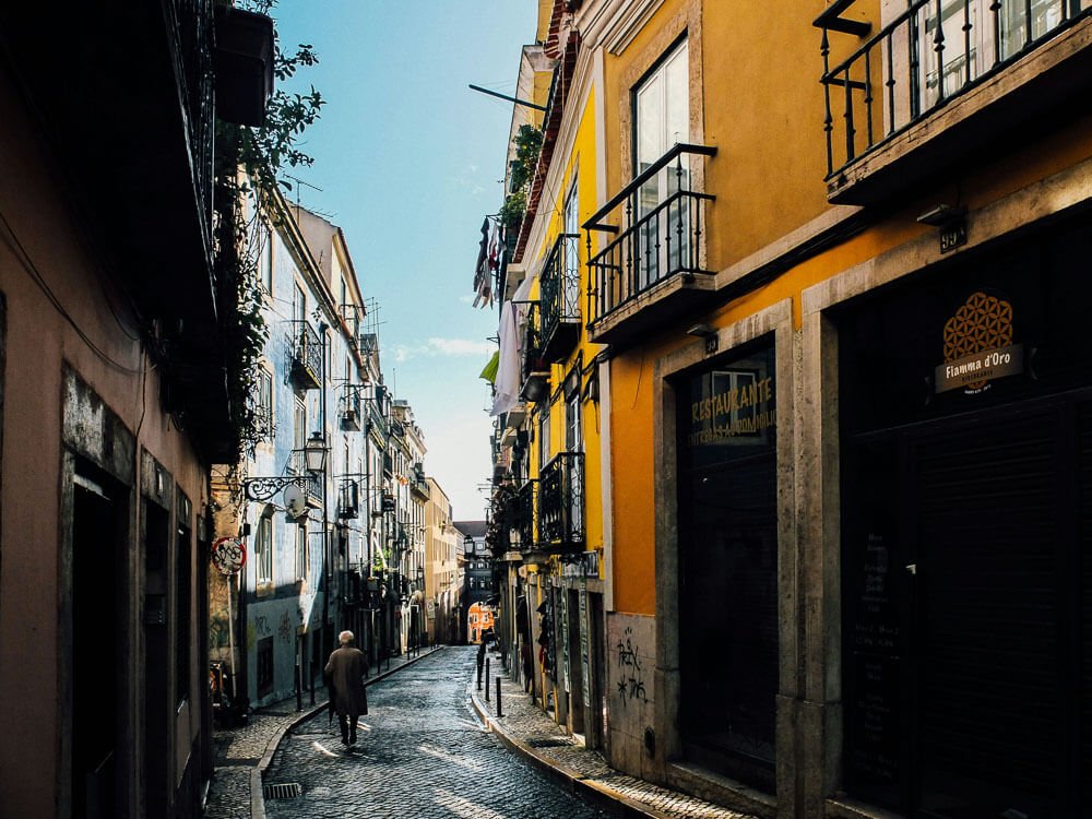 Street in the Bairro Alto, with cobblestone pavement and several buildings around with balconies. There is a person with their back to the photo.