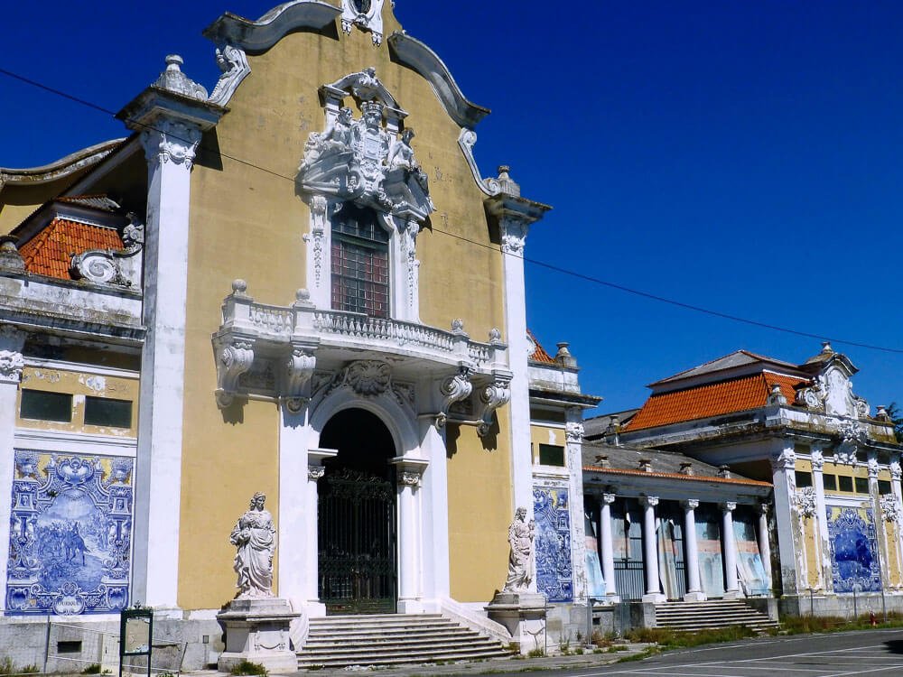 Entrance of the Pavilion, with an arched door, yellow walls, and white columns. There is a tile panel and a statue on each side.
