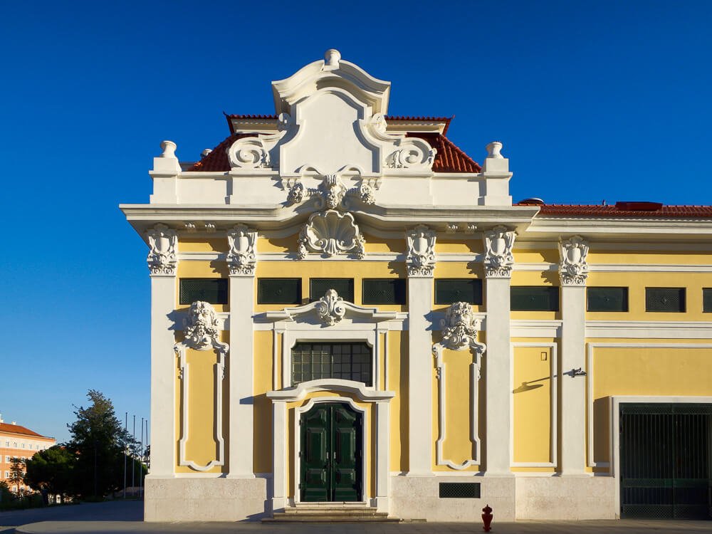 Facade of Pavilhão Carlos Lopes with a blue sky behind it. The building has yellow walls and ornate columns.
