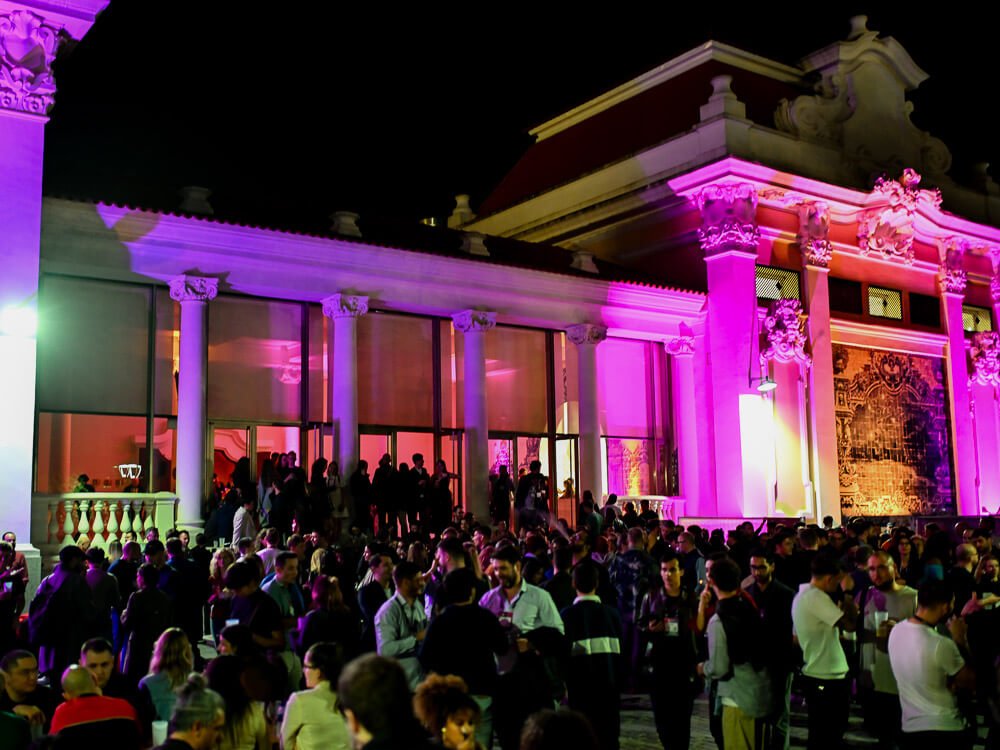 A crowd enjoying an event at Pavilhão Carlos Lopes during the night. There is pink light illuminating the columns and a large tile panel.

