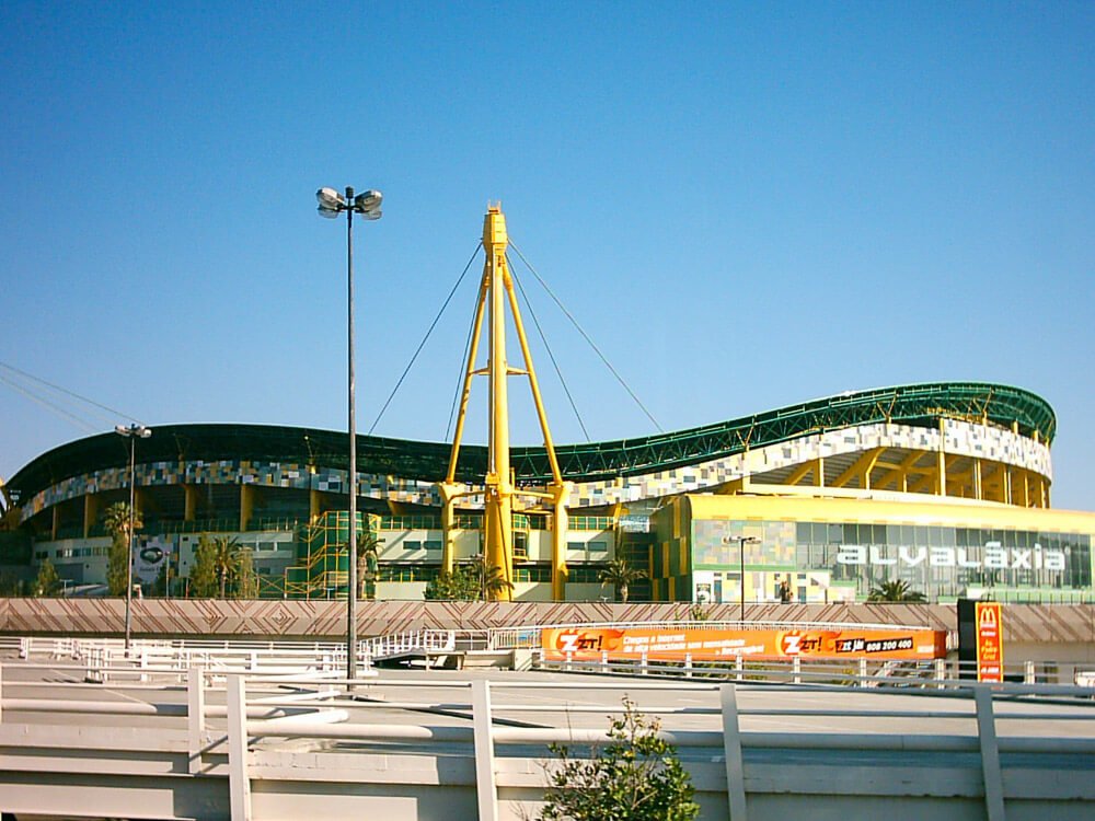 Estádio José Alvalade from the outside, with a blue sky behind it. There is a yellow steel structure, and the roof is undulating. The construction is yellow and green.