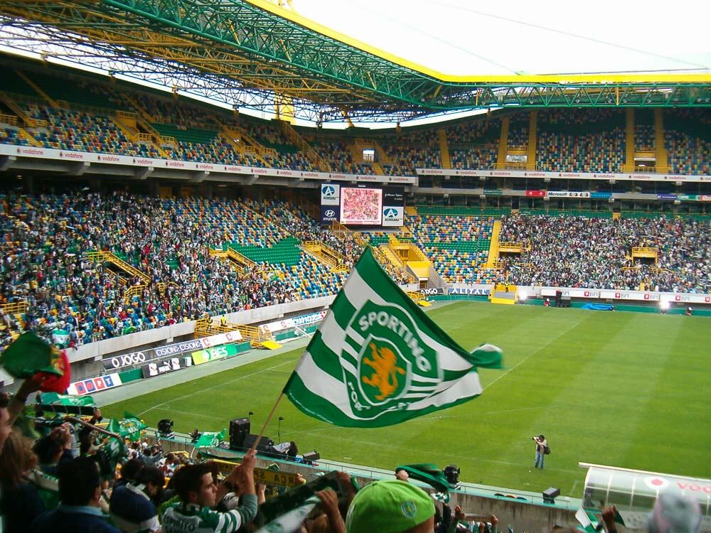 Stadium on a match day, with various fans in the stands. A man is waving a Sporting Portugal flag.