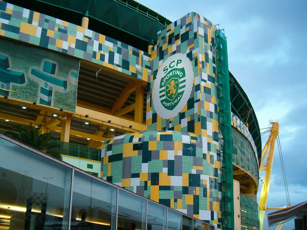 Detail of the façade of Estádio José Alvalade. There is a column with yellow, green, and gray tiles, and in the middle of it, the logo of Sporting Portugal.