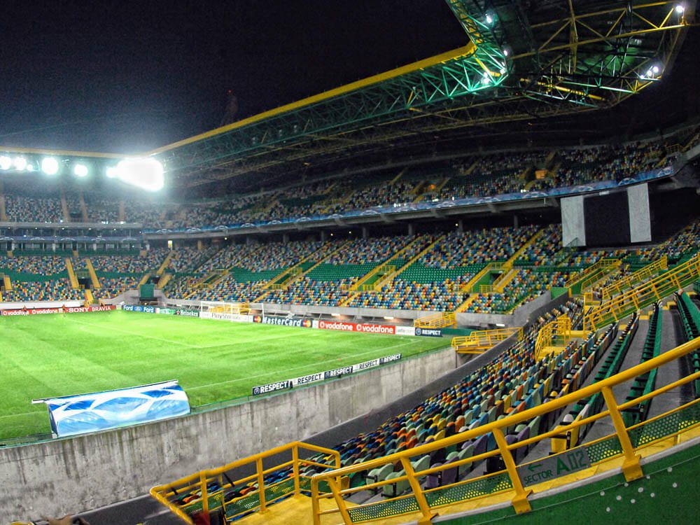 Interior of Estádio José Alvalade. In the center, a lawn. Around it, the seats, all colorful and separated by yellow handrails.