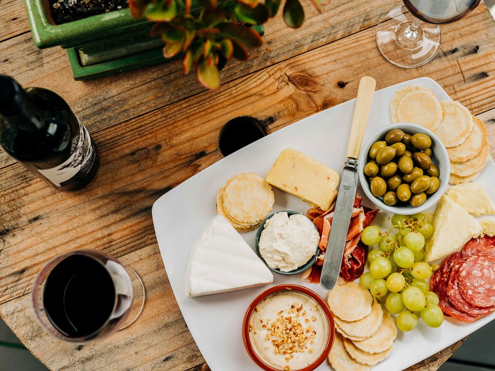 A board with cheeses, cold cuts, grapes, and olives on top of a wooden table. There is a bottle of wine and a full glass beside it. Above, there is a green plant vase.