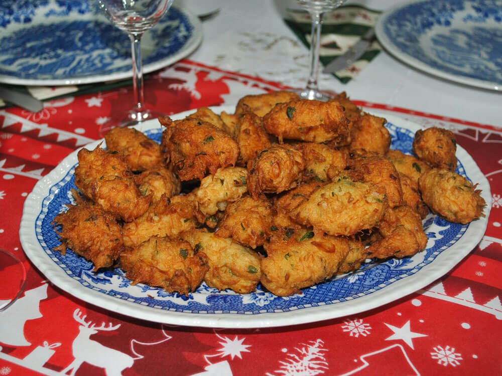 A blue and white plate full of codfish cakes. In the background, there is an empty plate and two empty glasses. The table is covered with a red Christmas-themed tablecloth.