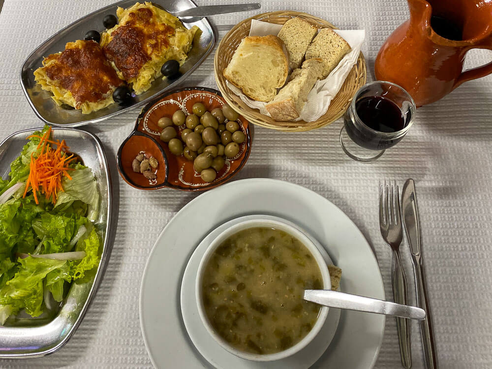 A dining table with a white tablecloth and various dishes on separate trays, a clay jug, and a glass of wine. On top of a white plate, there is a bowl of caldo verde soup.