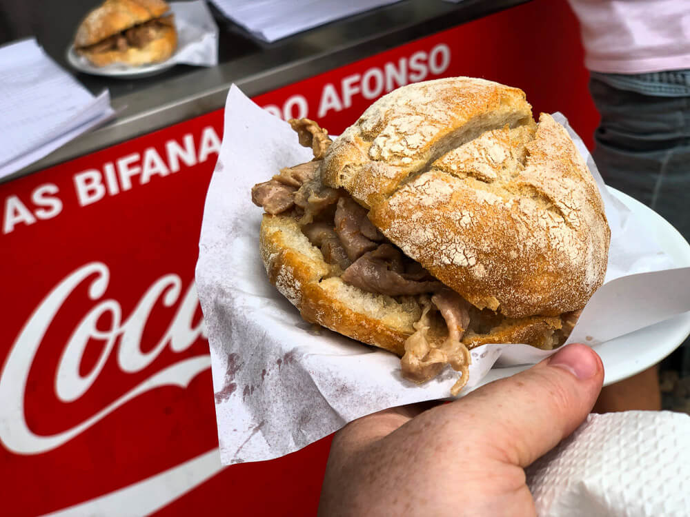 A person holding a Portuguese sandwich called bifana. Behind, there is a red Coca-Cola sign reading "bifanas do Afonso".