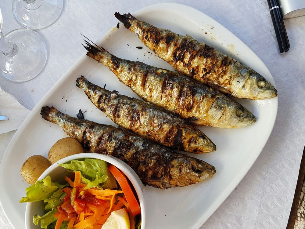 A white platter with four whole grilled sardines, next to it there is a bowl of salad and its potatoes.
