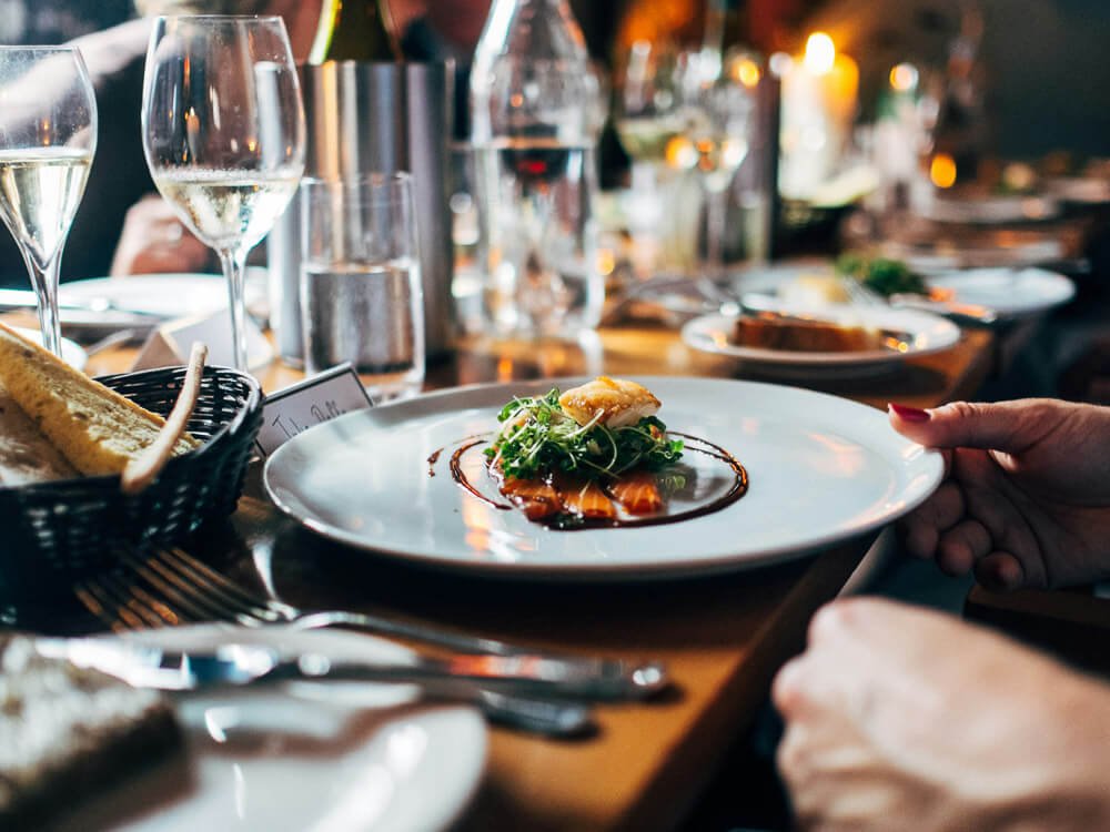 A restaurant table with various dishes served and wine glasses. One of the dishes and a bread basket are in focus, while the rest is blurred. There is a person with painted nails touching the plate.