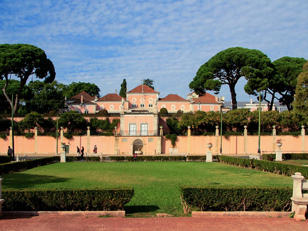 Facade of the Belém Palace, painted pink. There is a large grassy area in front and trees around it. Some people walk in front of the wall.