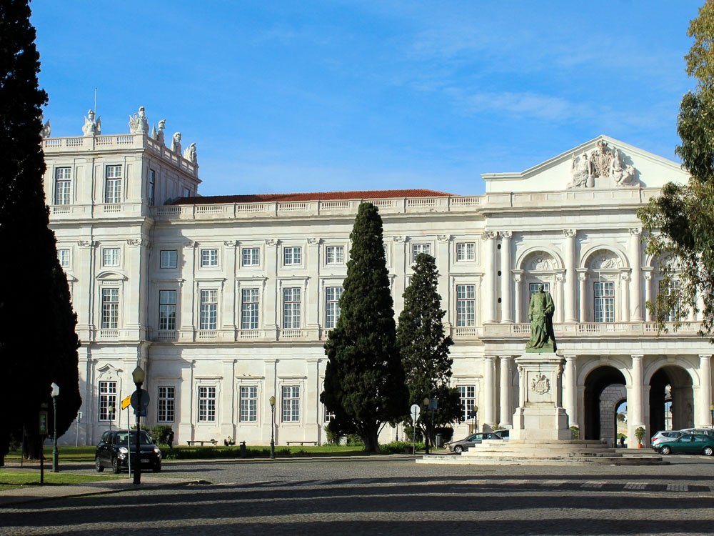 Facade of the National Palace of Ajuda. The walls are white, there is a roundabout with a statue in front, some cars, and trees.