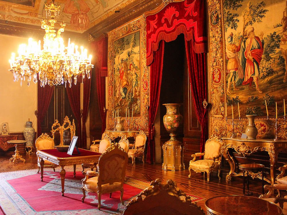 Interior of the National Palace of Ajuda. There are two large tapestries hanging on the wall, with a red curtain between them. There are various different pieces of furniture, such as a sideboard, table, and chairs. On the left side, there is a chandelier.