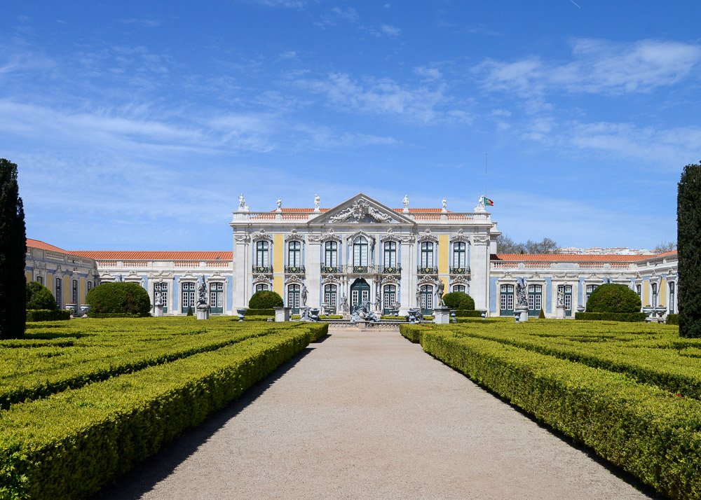 Facade of the Queluz Palace, with white, blue, and yellow walls. There are trimmed bushes on both sides of a concrete path leading to the palace, and statues in front of it.