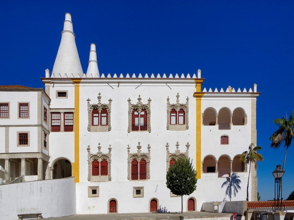 Facade of the National Palace of Sintra. The walls are white, with red windows framed with tower-shaped details. There are two towers on the left side, and some trees on the right side.