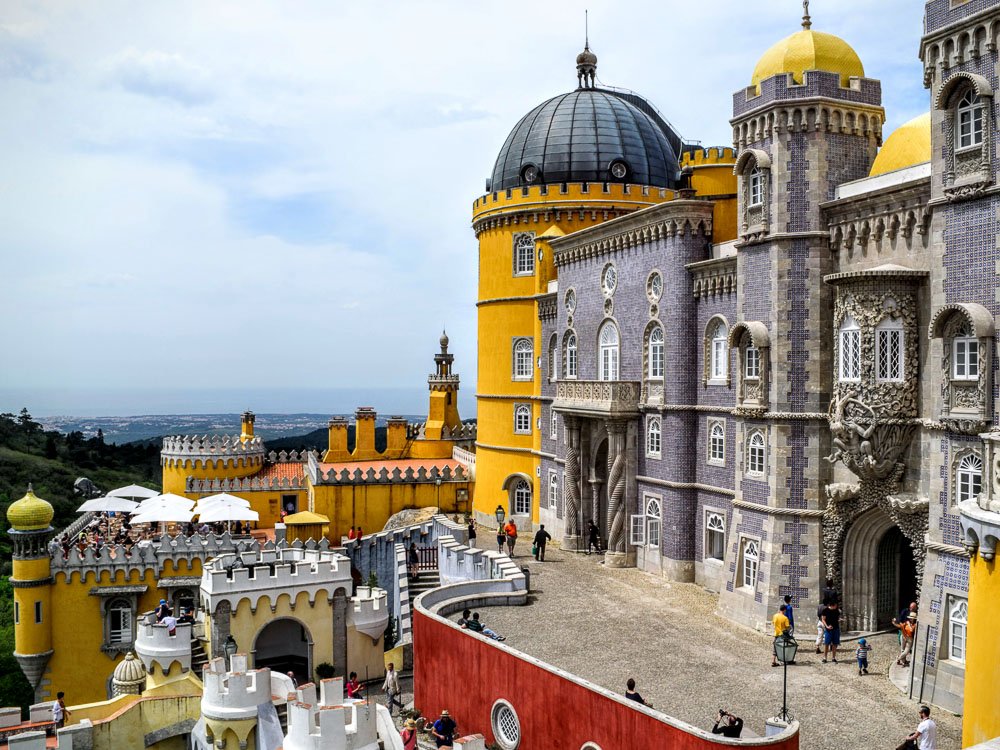 View of the National Palace of Pena. On the right side, a building with tiles. Behind it, a yellow building with a dome. There are people walking, and in the background, trees.