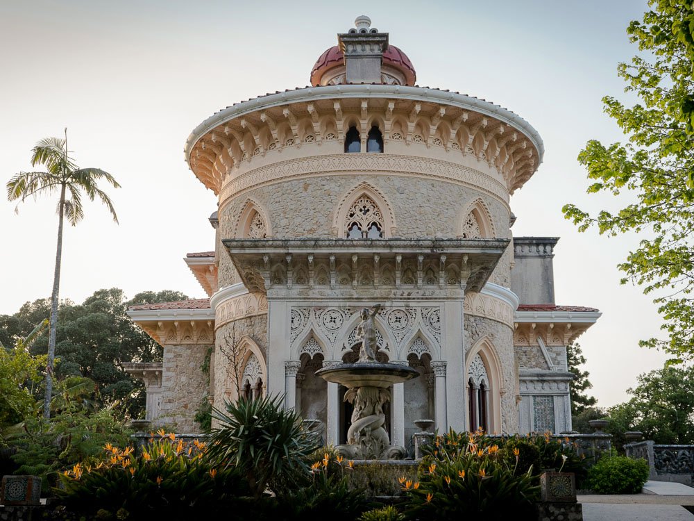 Facade of the Monserrate Palace. A round building, which becomes rectangular further back, framed by trees. In front, there is a fountain with statues and a flowerbed.