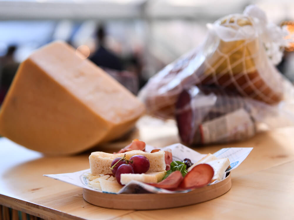 A wooden table with a plate of bread, grapes, and Portuguese sausage. In the background, out of focus, a triangular piece of cheese and a piece of packaged cured ham.