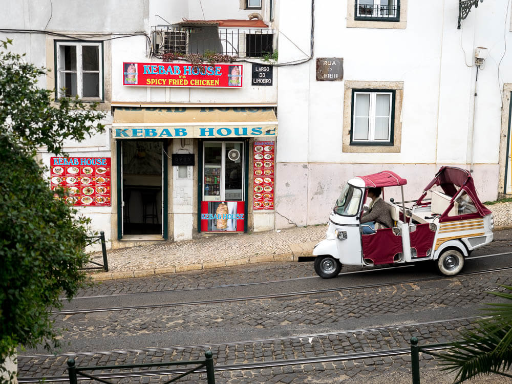 A red and white tuk-tuk parked on a cobblestone street in Lisbon. On the left side, there is a kebab restaurant.