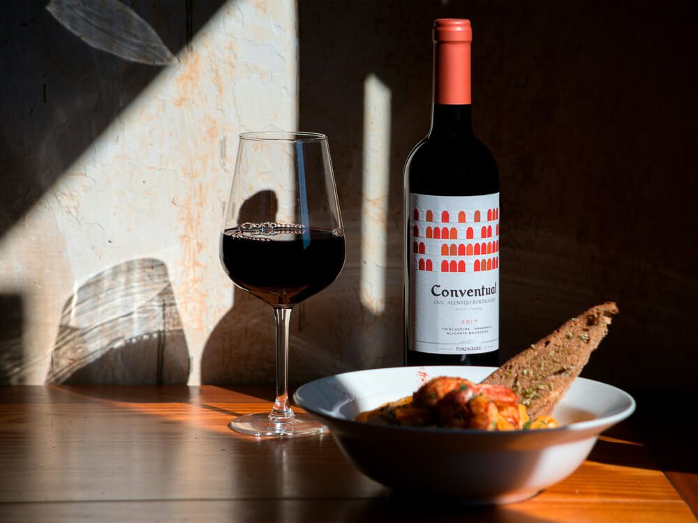 A wooden table with a deep dish of food and a glass of red wine, with a wine bottle beside it.