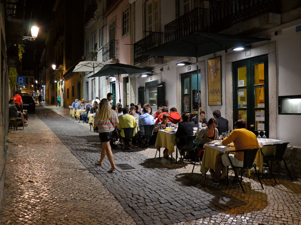 Diners enjoying a lively evening at an outdoor restaurant on a cobblestone street in Lisbon, with tables set along the sidewalk and people walking by.