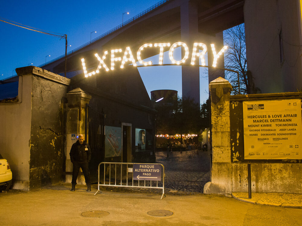 The illuminated entrance to LX Factory at dusk, showcasing the vibrant cultural and creative hub under the 25 de Abril Bridge in Lisbon.