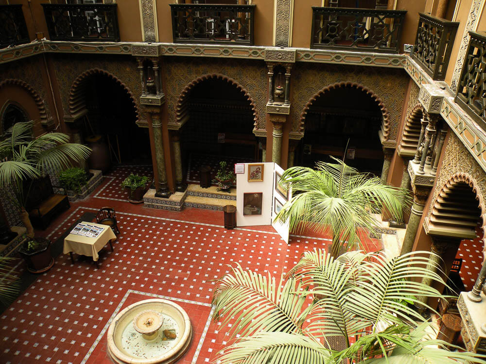 An ornate courtyard with arched columns, tiled floors, and potted plants, featuring a central fountain and art displays.