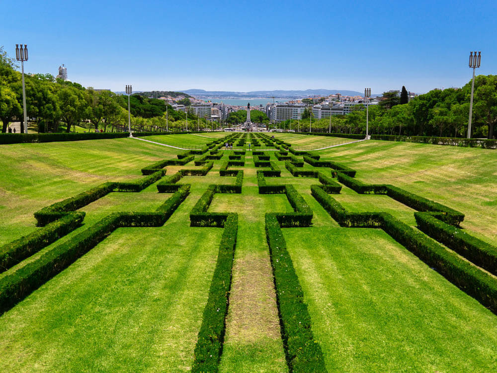 A view from the top of Eduardo VII Park in Lisbon, looking down the meticulously manicured hedges towards the cityscape and Tagus River in the distance.