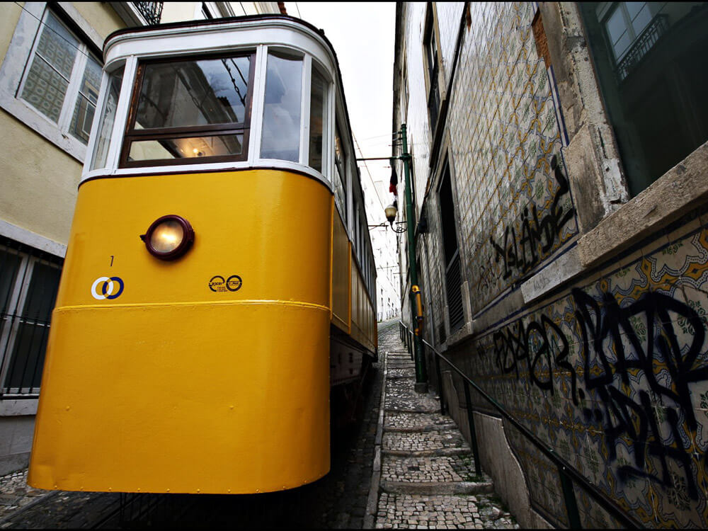 A bright yellow tram navigating a narrow, graffiti-covered street with tiled walls.