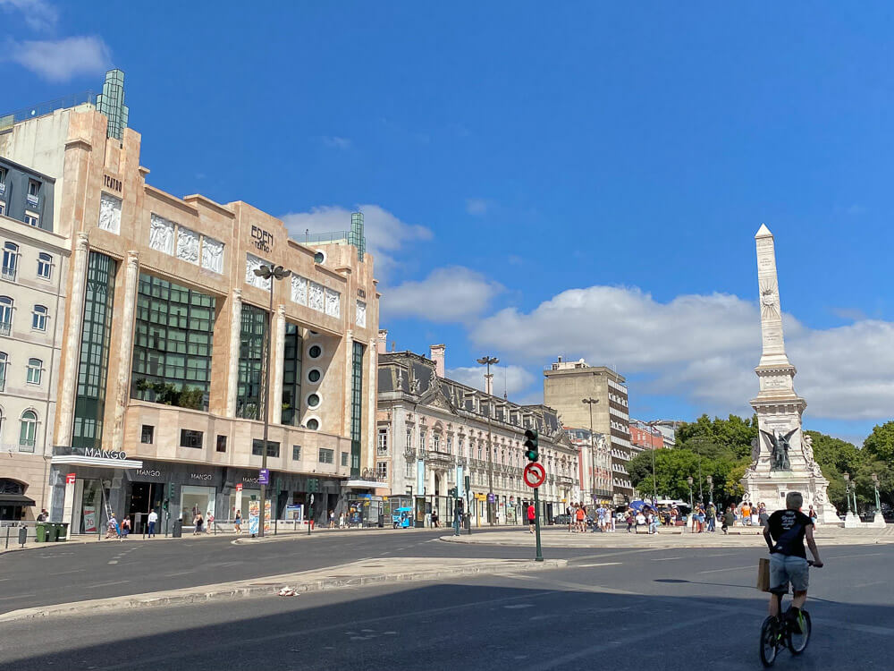 A cyclist rides past the Monument to the Restorers in Lisbon, with the iconic art deco Eden Theatre building in the background under a bright blue sky.