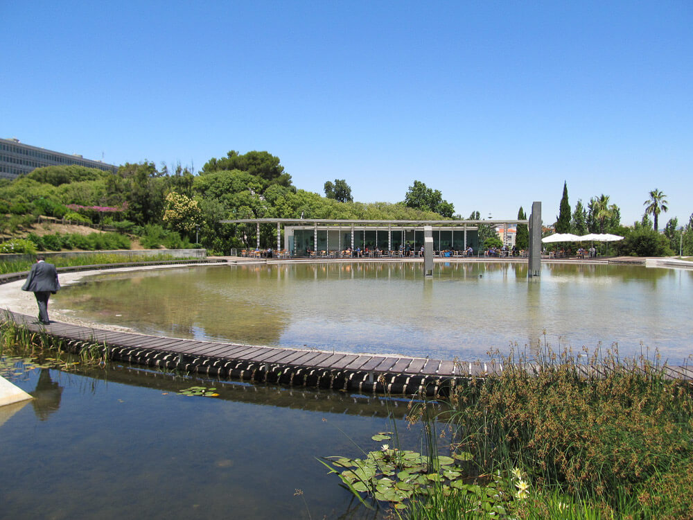 A pond with a wooden walkway surrounded by greenery and a modern café in the background.